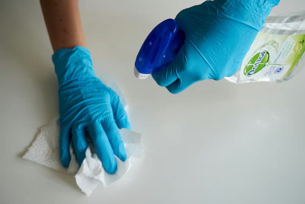 Person in blue gloves cleaning a surface with spray cleaner and paper towel.
